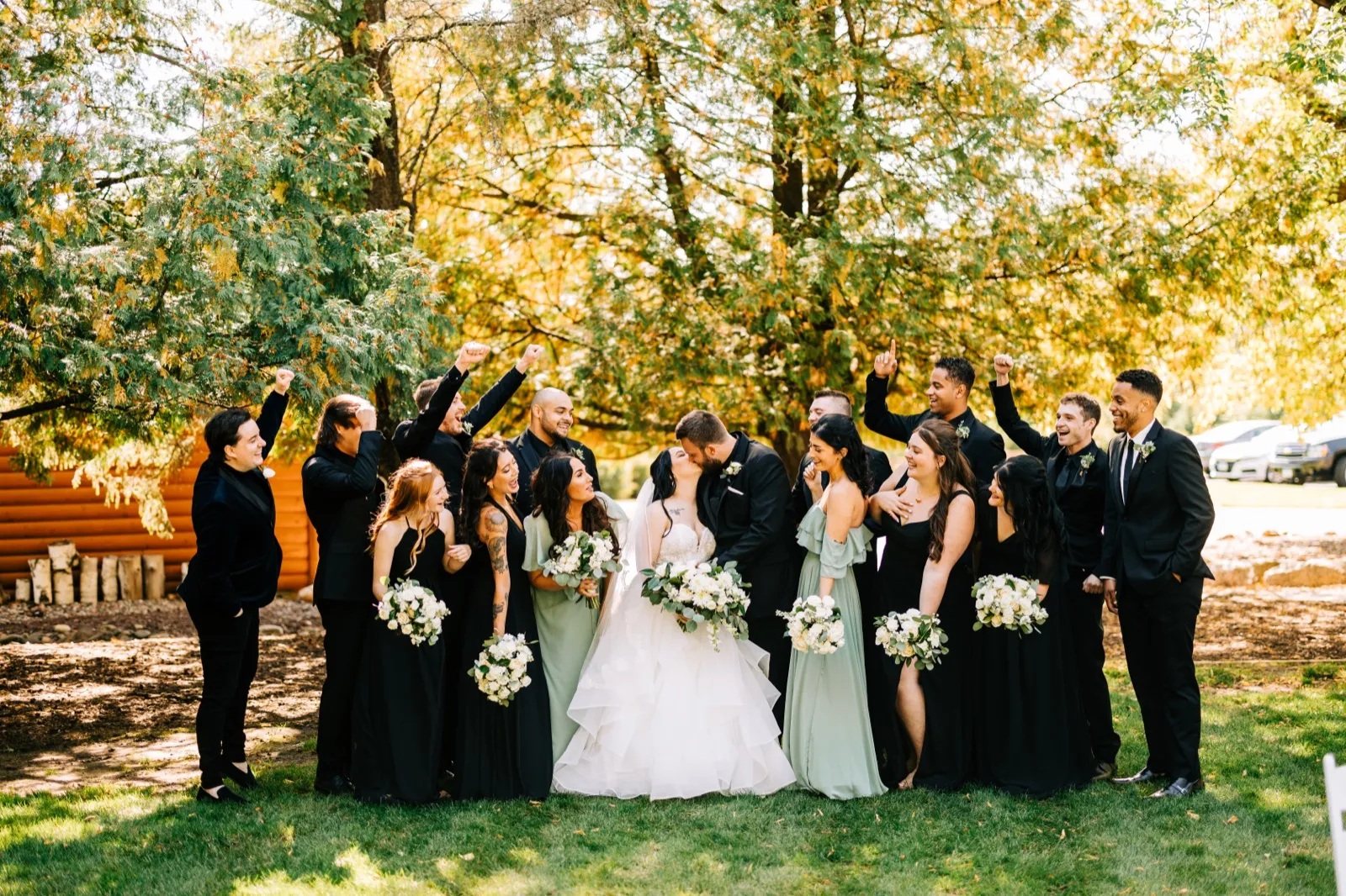 The couple kisses at the center of their bridal party while groomsmen and bridesmaids cheer with raised fists on a sunlit lawn beside a log cabin — Tim Larsen Photography, Brainerd Lakes MN