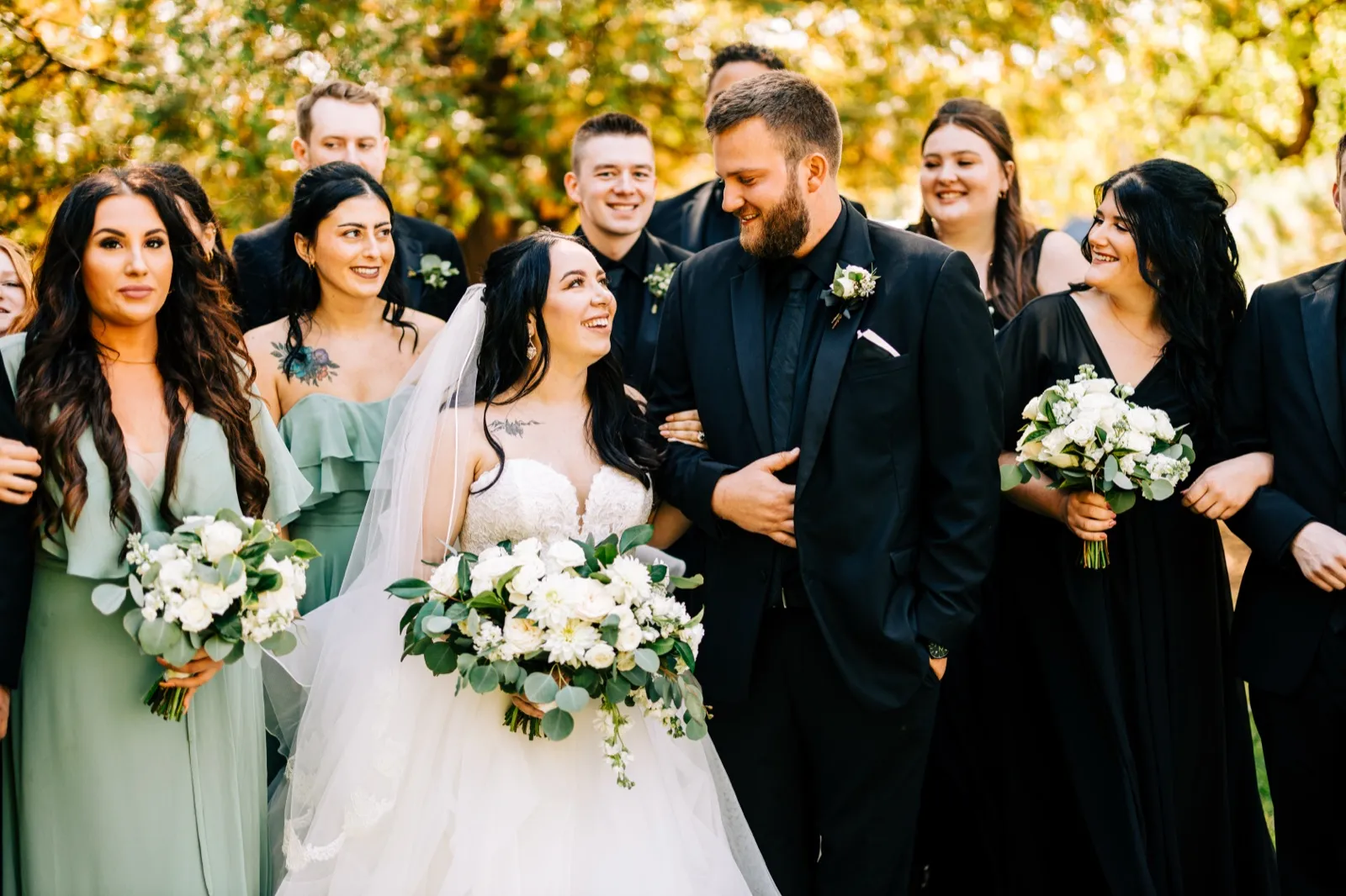 The bride and groom share a laugh together as their bridal party stands beside them in sage and black attire, framed by warm autumn foliage — Tim Larsen Photography, Brainerd Lakes MN