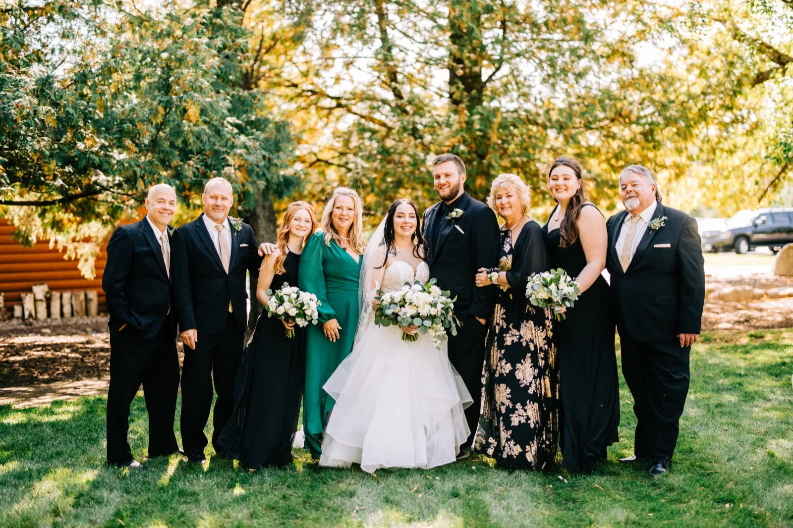 A wedding party of nine, including the bride and groom flanked by family members in black and emerald attire, stands together on a grass lawn beneath a large backlit tree — Tim Larsen Photography, Brainerd Lakes MN