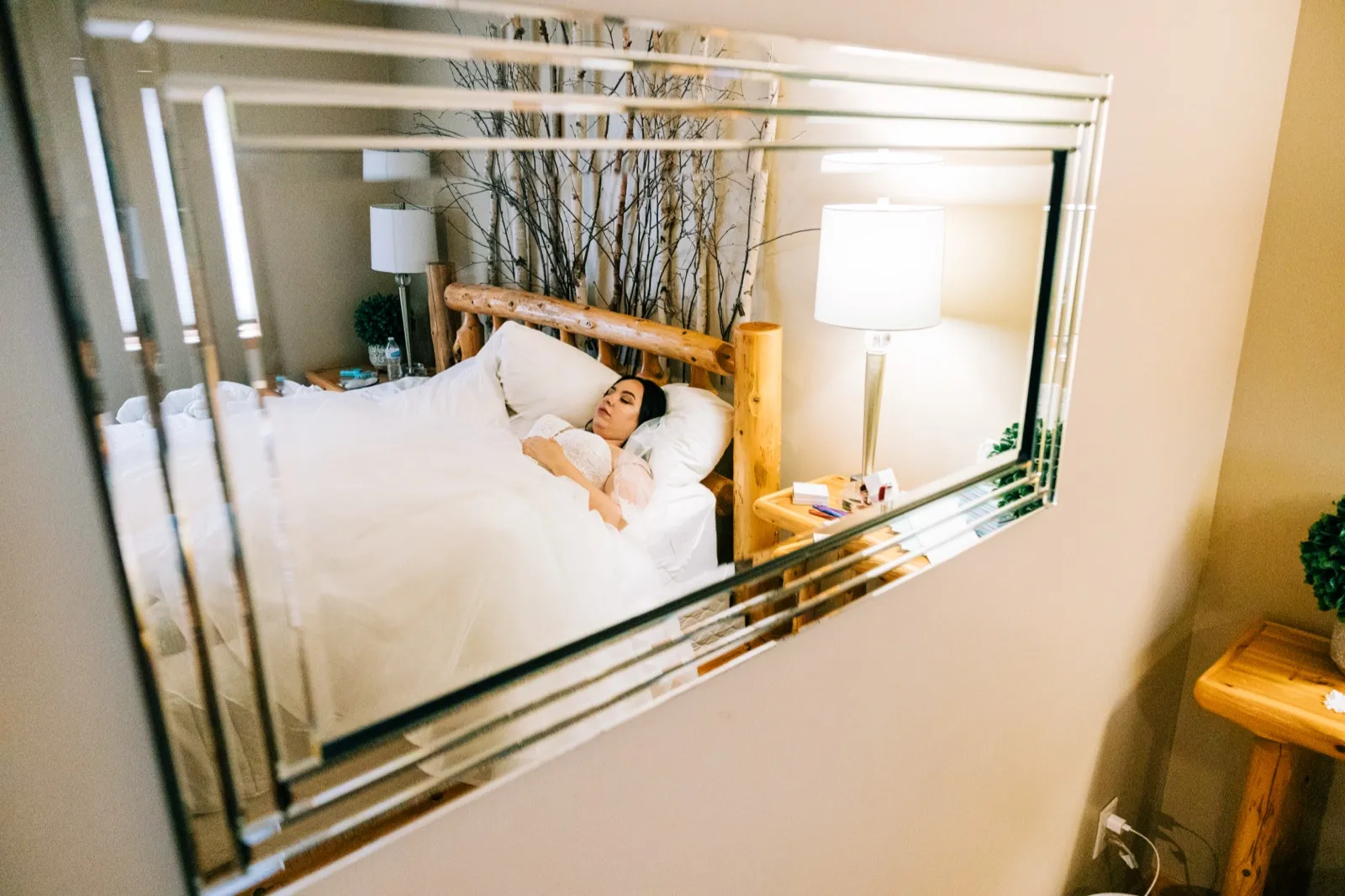 The bride rests on a log-frame bed in a cabin bridal suite, seen through the reflection of a decorative mirror with layered glass edges — Tim Larsen Photography, Brainerd Lakes MN