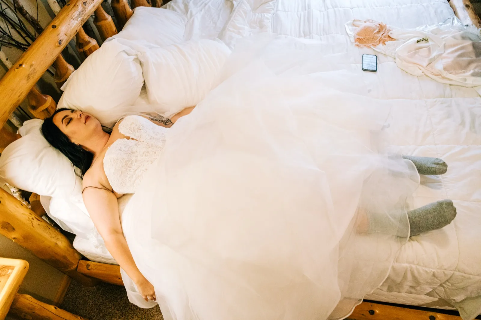 The bride lies back on a log-frame bed in her wedding gown with eyes closed, her tulle skirt and veil spilling across the white bedding next to a phone and discarded items — Tim Larsen Photography, Brainerd Lakes MN
