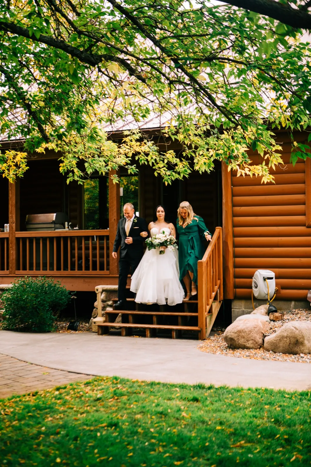 The bride descends a wooden cabin staircase flanked by her father and mother, carrying a white bouquet, framed by leafy tree branches overhead — Tim Larsen Photography, Brainerd Lakes MN