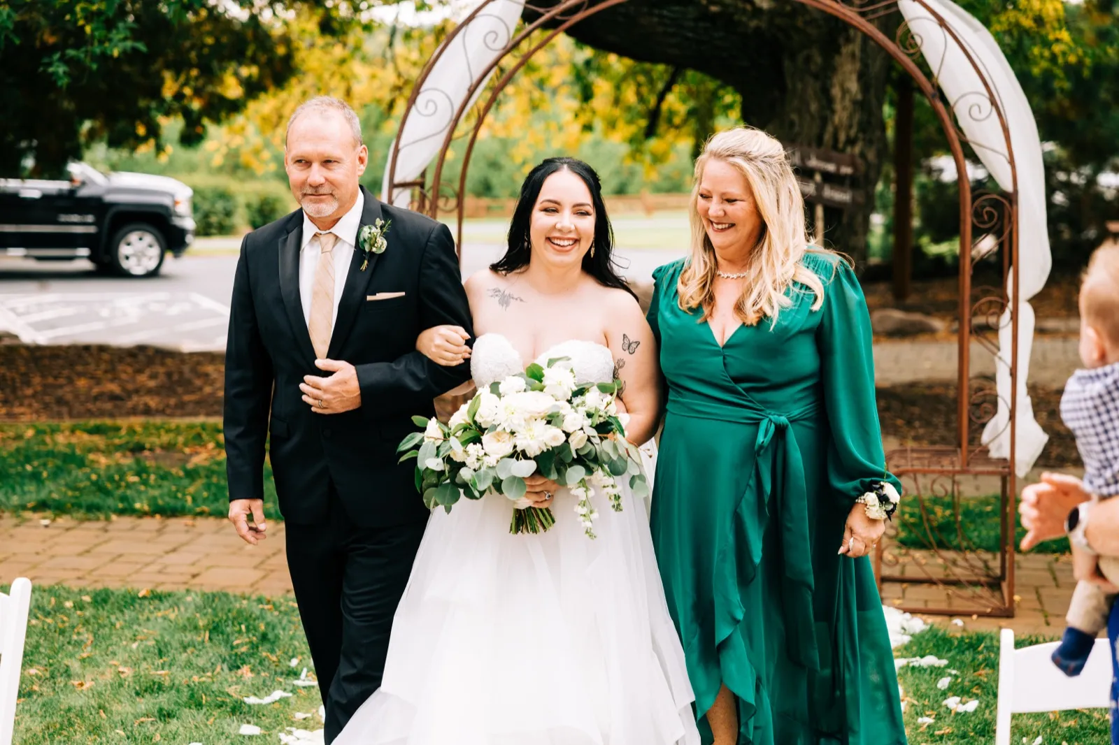 The bride walks down the aisle between her parents, smiling broadly while holding a large white bouquet, beneath a wrought-iron ceremony arch — Tim Larsen Photography, Brainerd Lakes MN