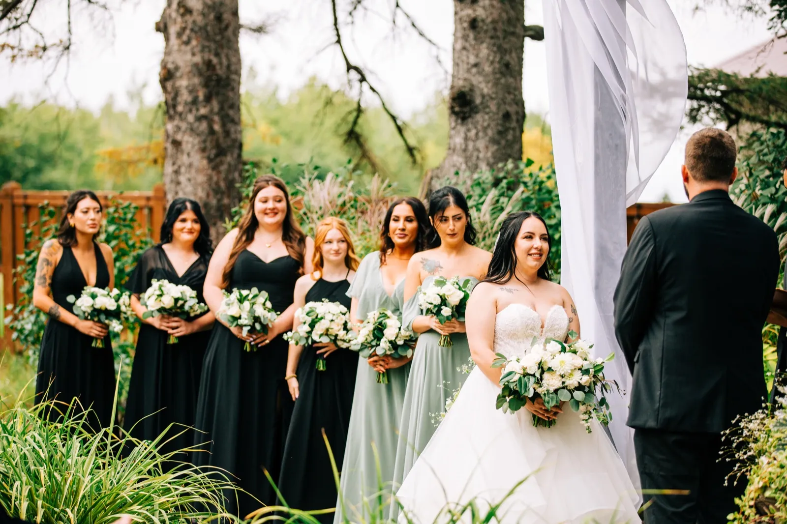 The bride and her bridesmaids stand in a row holding white bouquets at an outdoor ceremony, facing the groom beside a billowing white drape — Tim Larsen Photography, Brainerd Lakes MN