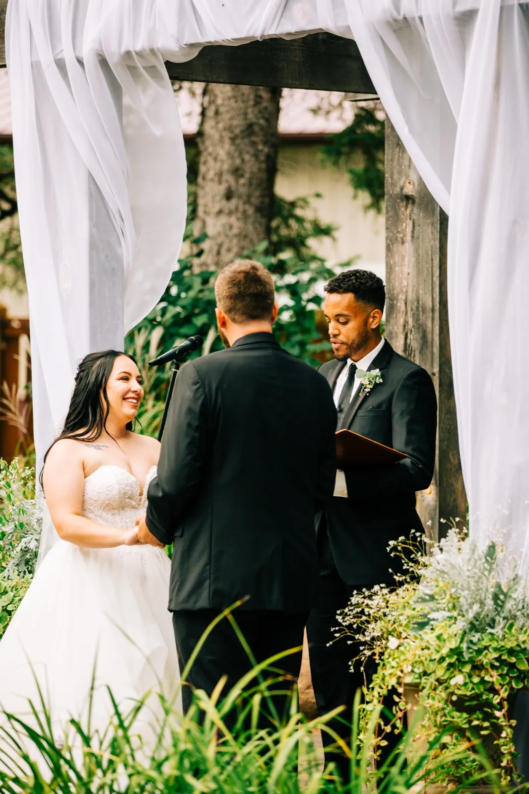 The bride smiles at her groom while the officiant reads from a book beside them under a draped wooden ceremony arch surrounded by greenery — Tim Larsen Photography, Brainerd Lakes MN