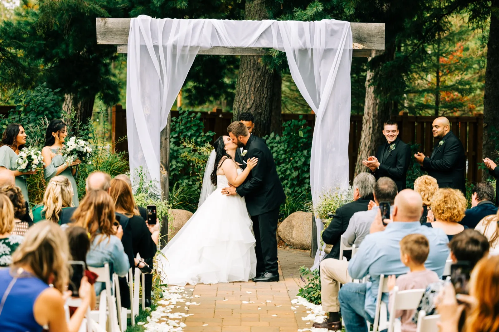 The bride and groom share their first kiss beneath a wooden arch draped in white fabric while bridal party and guests applaud in a forested ceremony space — Tim Larsen Photography, Brainerd Lakes MN