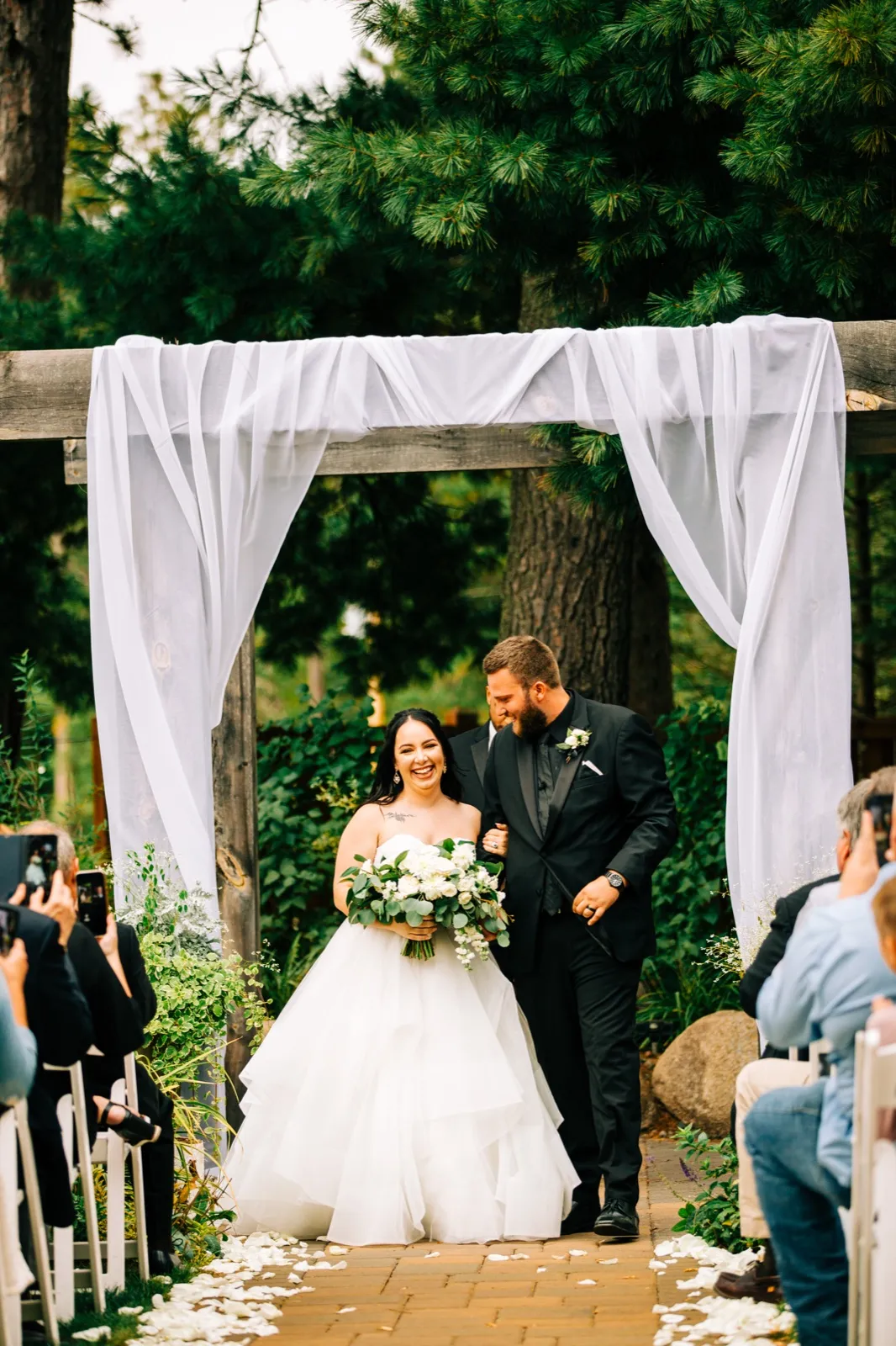 The bride beams holding a white bouquet as she walks down the brick aisle with the groom beneath a draped wooden arch framed by tall pines at Pine Peaks Event Center — Tim Larsen Photography, Brainerd Lakes MN