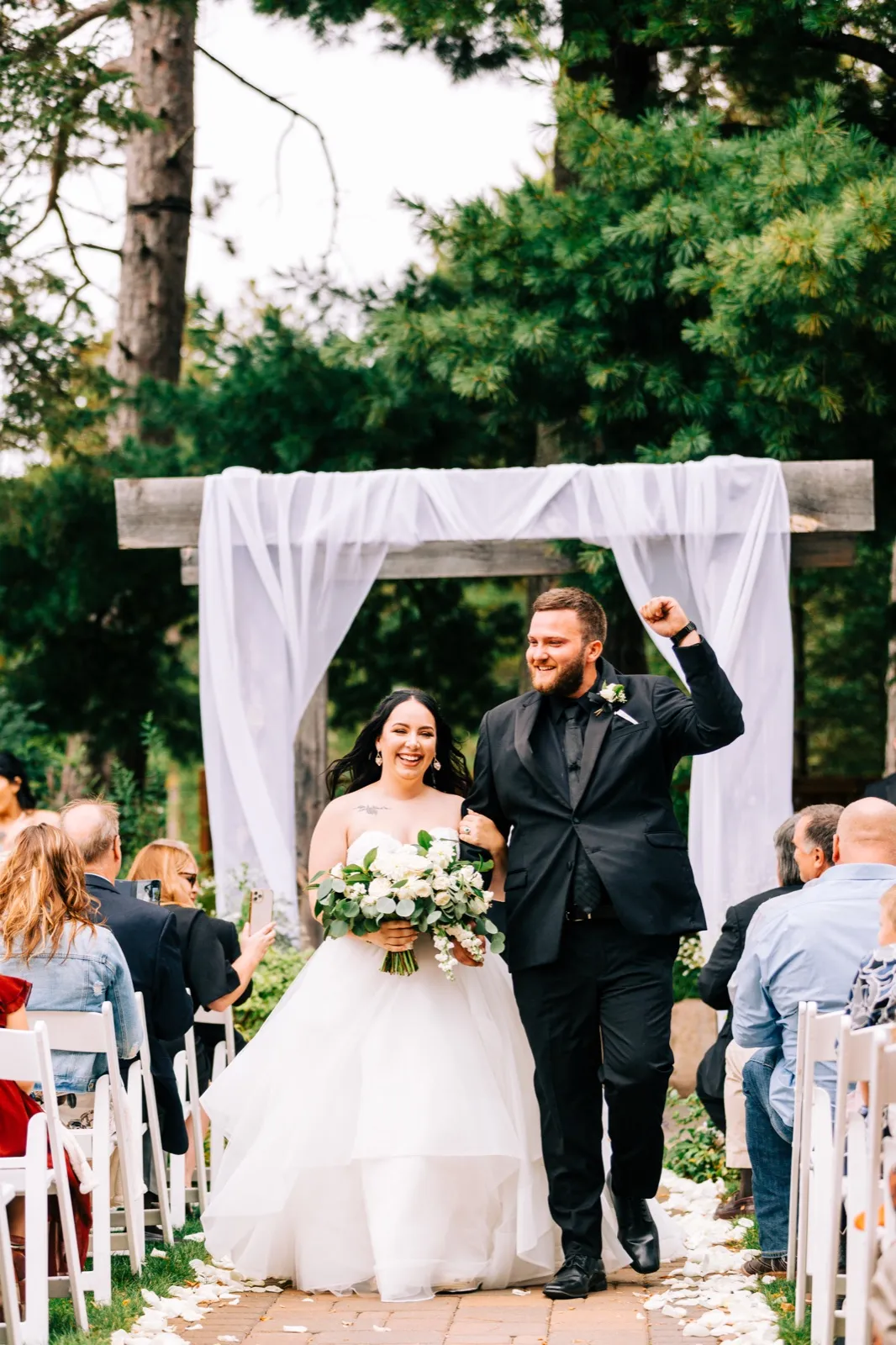 The bride beams holding her white bouquet while the groom raises a triumphant fist as they walk back down the aisle beneath a draped wooden arch surrounded by tall pines — Tim Larsen Photography, Brainerd Lakes MN