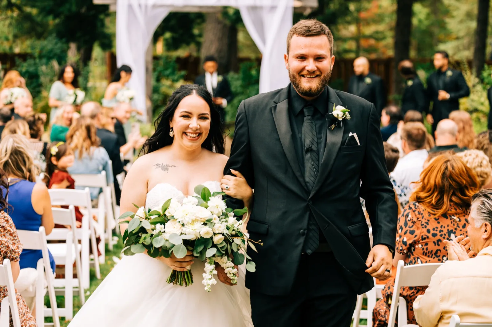 Taylor laughs broadly while holding a large white bouquet as she walks down the aisle arm-in-arm with Charles, surrounded by seated guests at an outdoor forest ceremony. — Tim Larsen Photography, Brainerd Lakes MN