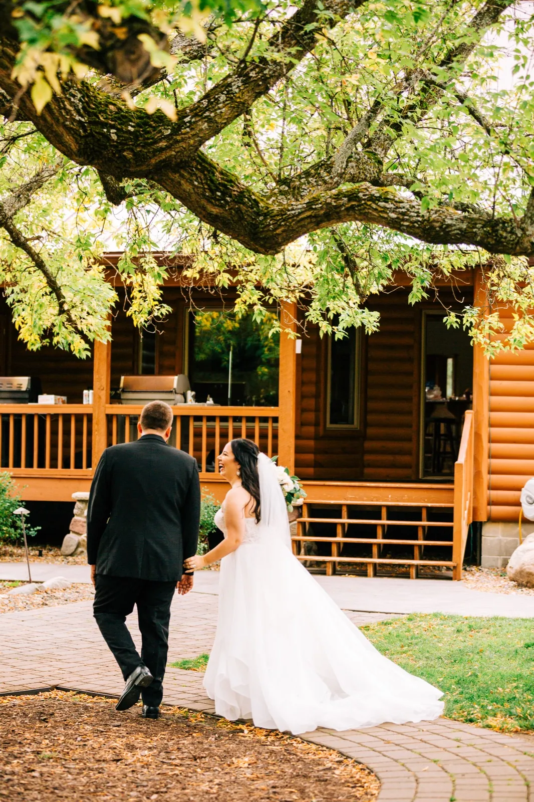 Taylor laughs while holding Charles's hand as they walk together beneath a sprawling tree toward a log cabin porch at Pine Peaks Event Center. — Tim Larsen Photography, Brainerd Lakes MN