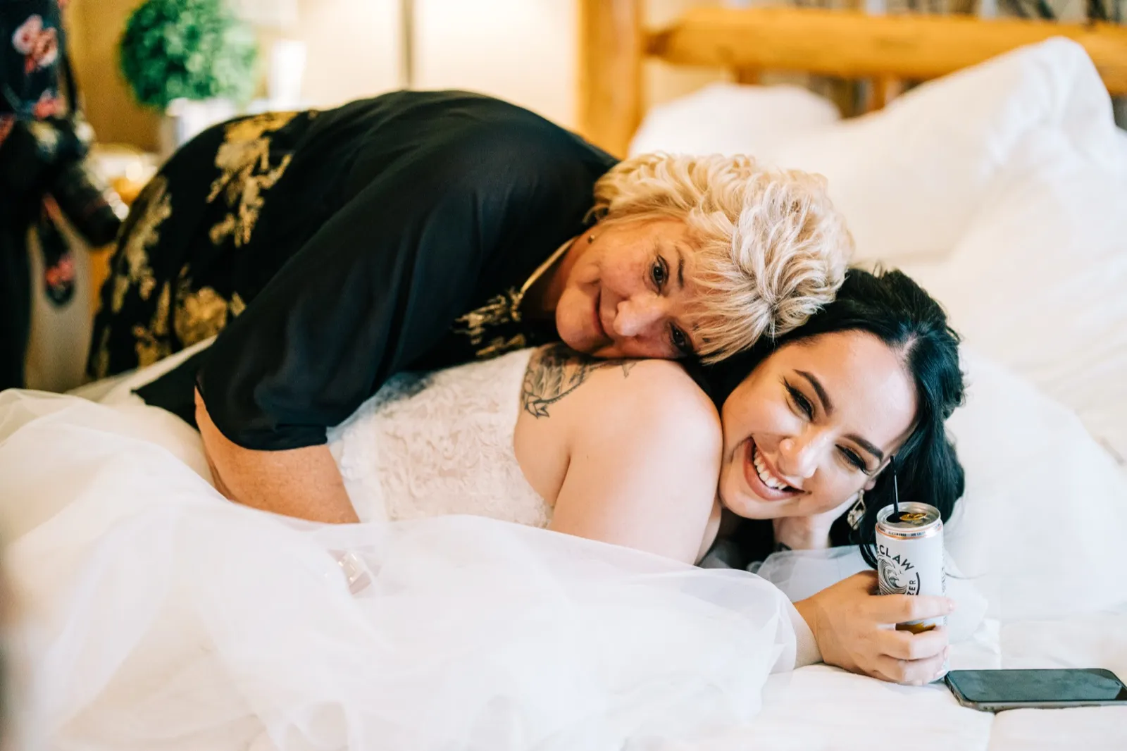 Taylor laughs as Charles's mother rests her head on her shoulder while they lie together on a white bed in the bridal suite. — Tim Larsen Photography, Brainerd Lakes MN