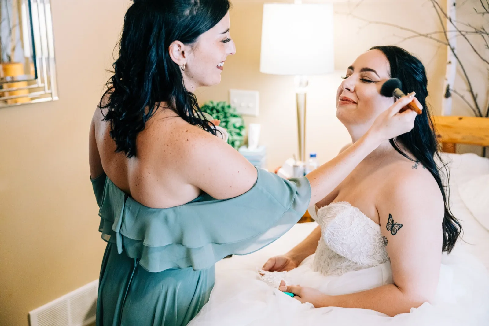 A bridesmaid in a sage off-the-shoulder dress touches up Taylor's blush as she sits on the bed in her wedding gown. — Tim Larsen Photography, Brainerd Lakes MN