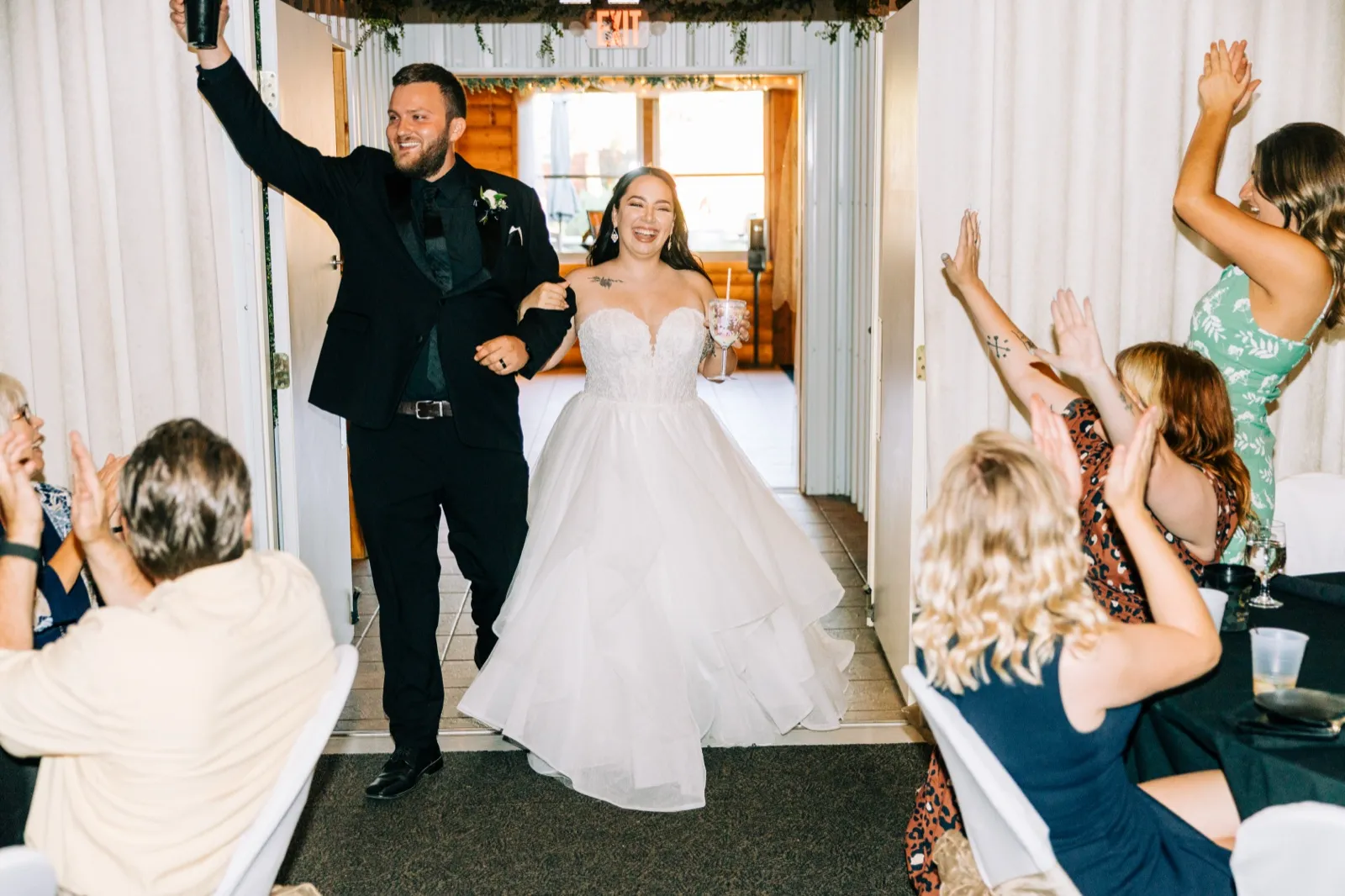 The groom raises a drink overhead and the bride laughs broadly as they enter the reception through a curtained doorway flanked by cheering guests with raised arms — Tim Larsen Photography, Brainerd Lakes MN