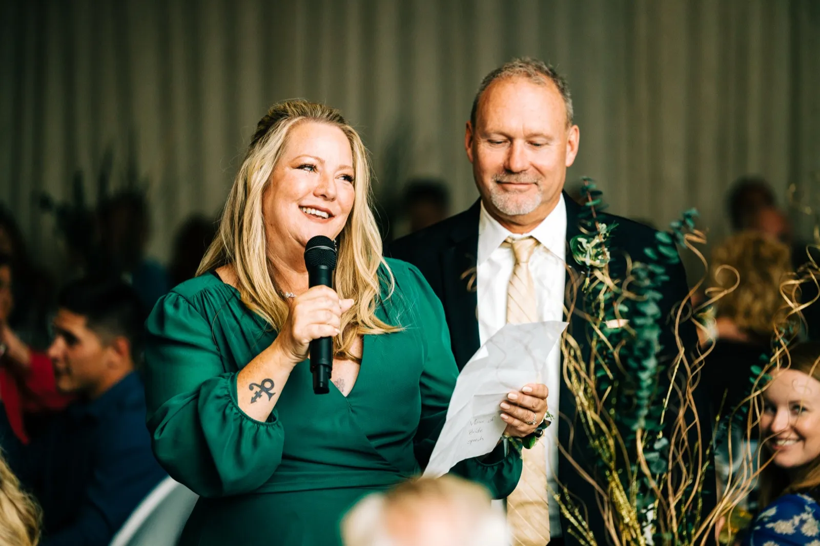 A woman in an emerald green dress laughs while speaking into a microphone, standing beside a man in a dark suit reading from a folded paper — Tim Larsen Photography, Brainerd Lakes MN