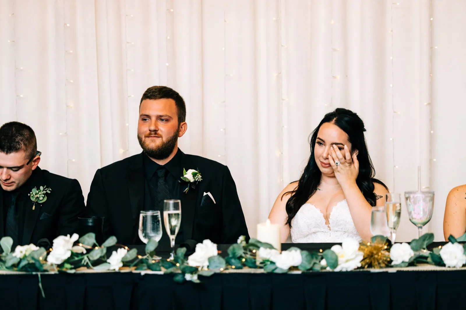 The bride wipes a tear at the head table while the groom listens intently during reception toasts, with greenery and champagne flutes in the foreground — Tim Larsen Photography, Brainerd Lakes MN