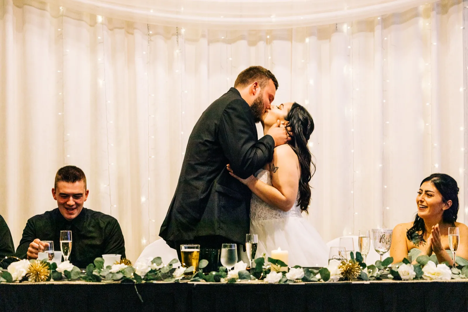 Charles holds Taylor's face as they kiss at the head table while a groomsman and bridesmaid laugh and clap on either side. — Tim Larsen Photography, Brainerd Lakes MN