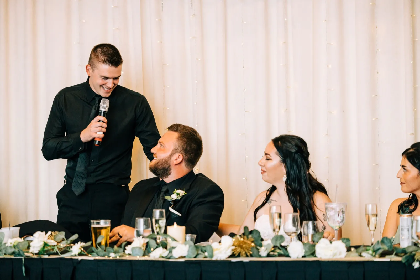 A best man holds a microphone mid-toast as Charles looks up smiling, with Taylor seated beside them at the floral-lined head table. — Tim Larsen Photography, Brainerd Lakes MN