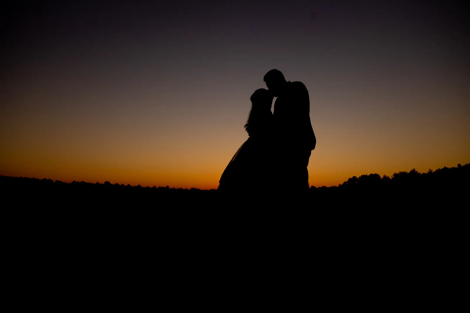 The bride and groom stand in silhouette against an orange-to-indigo dusk sky, foreheads tilted together above a dark treeline — Tim Larsen Photography, Brainerd Lakes MN