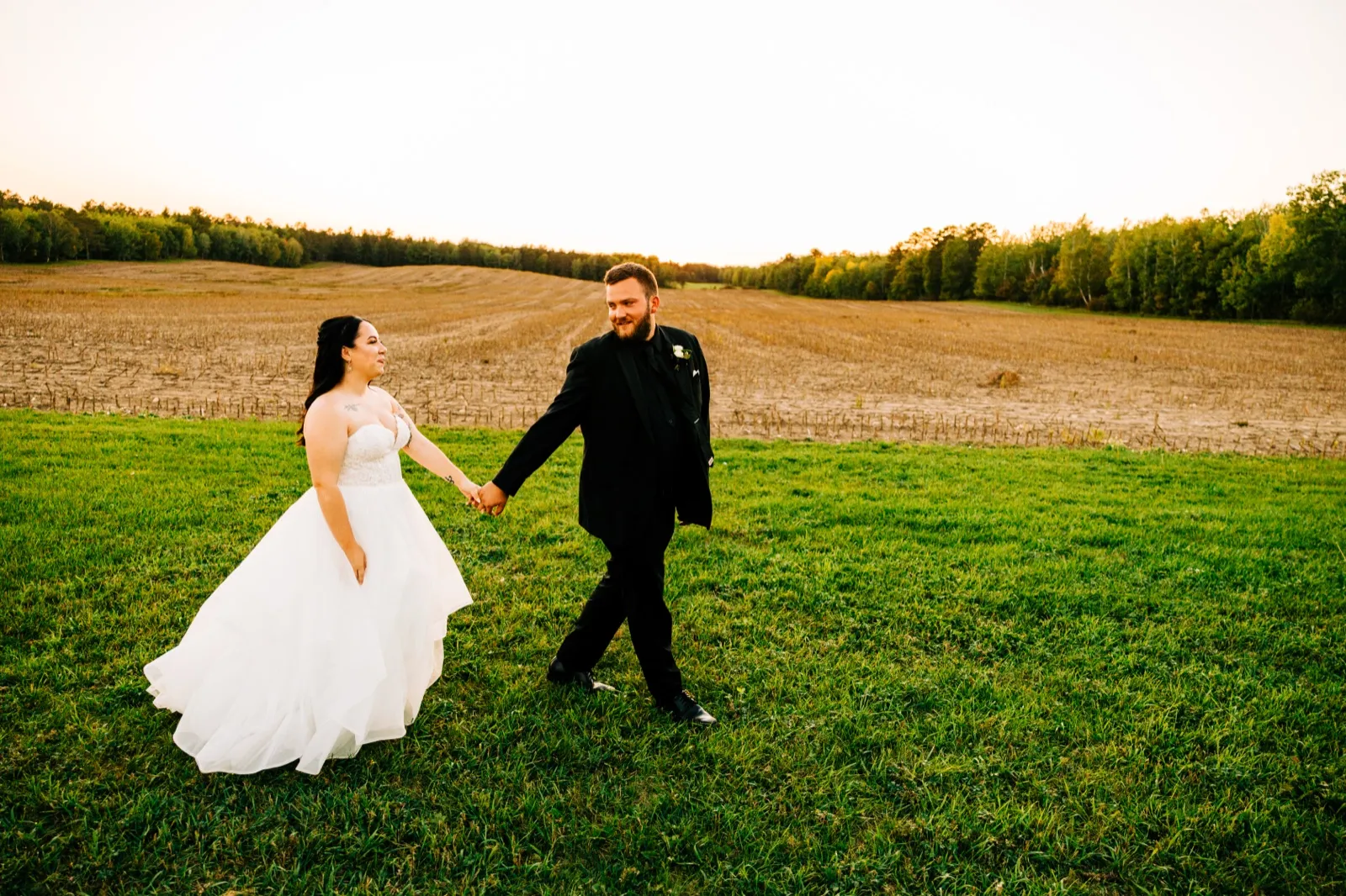 The bride in a full white gown is led by the groom in a black suit across a grassy field with a harvested cornfield and tree line behind them at sunset — Tim Larsen Photography, Brainerd Lakes MN
