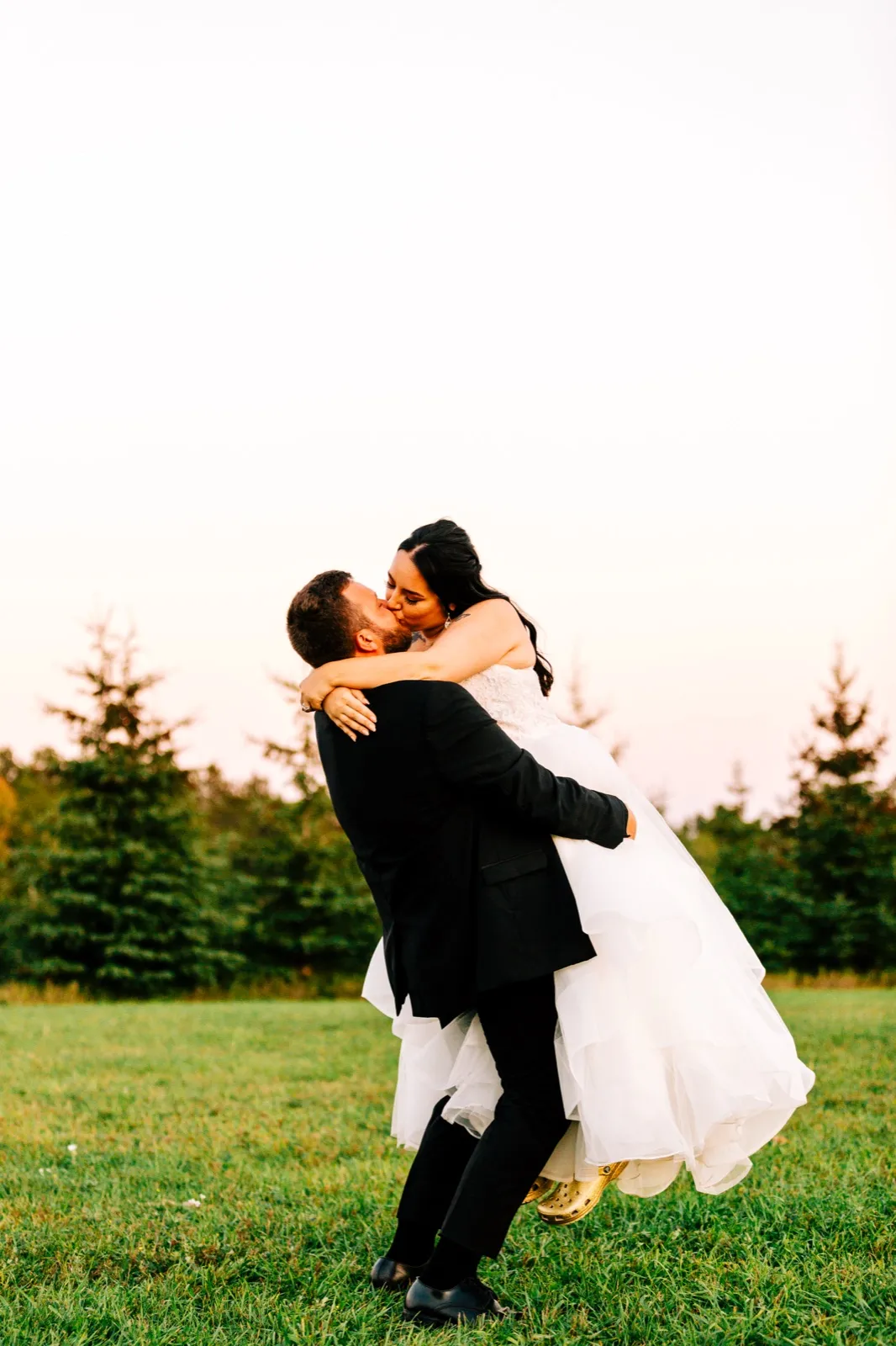 Charles lifts Taylor off the ground as they kiss in an open field at dusk, her gown spilling out around them with evergreen trees behind. — Tim Larsen Photography, Brainerd Lakes MN
