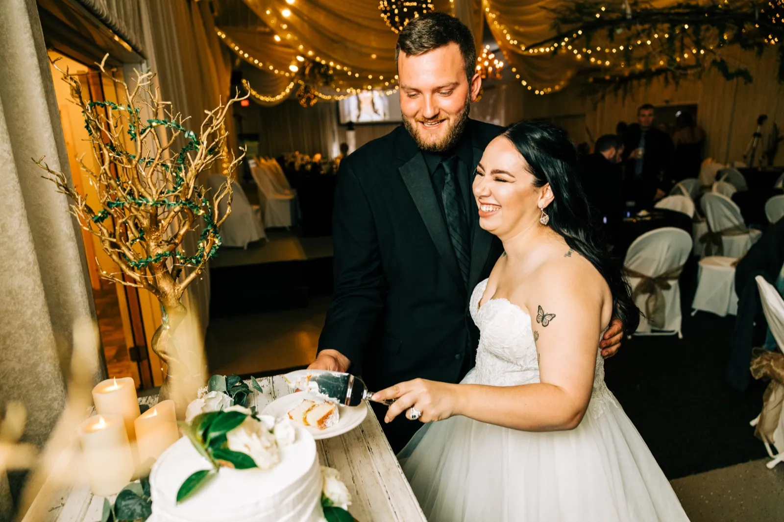 Taylor laughs with her eyes closed as Charles smiles down at her while they cut the cake together beneath a canopy of string lights. — Tim Larsen Photography, Brainerd Lakes MN