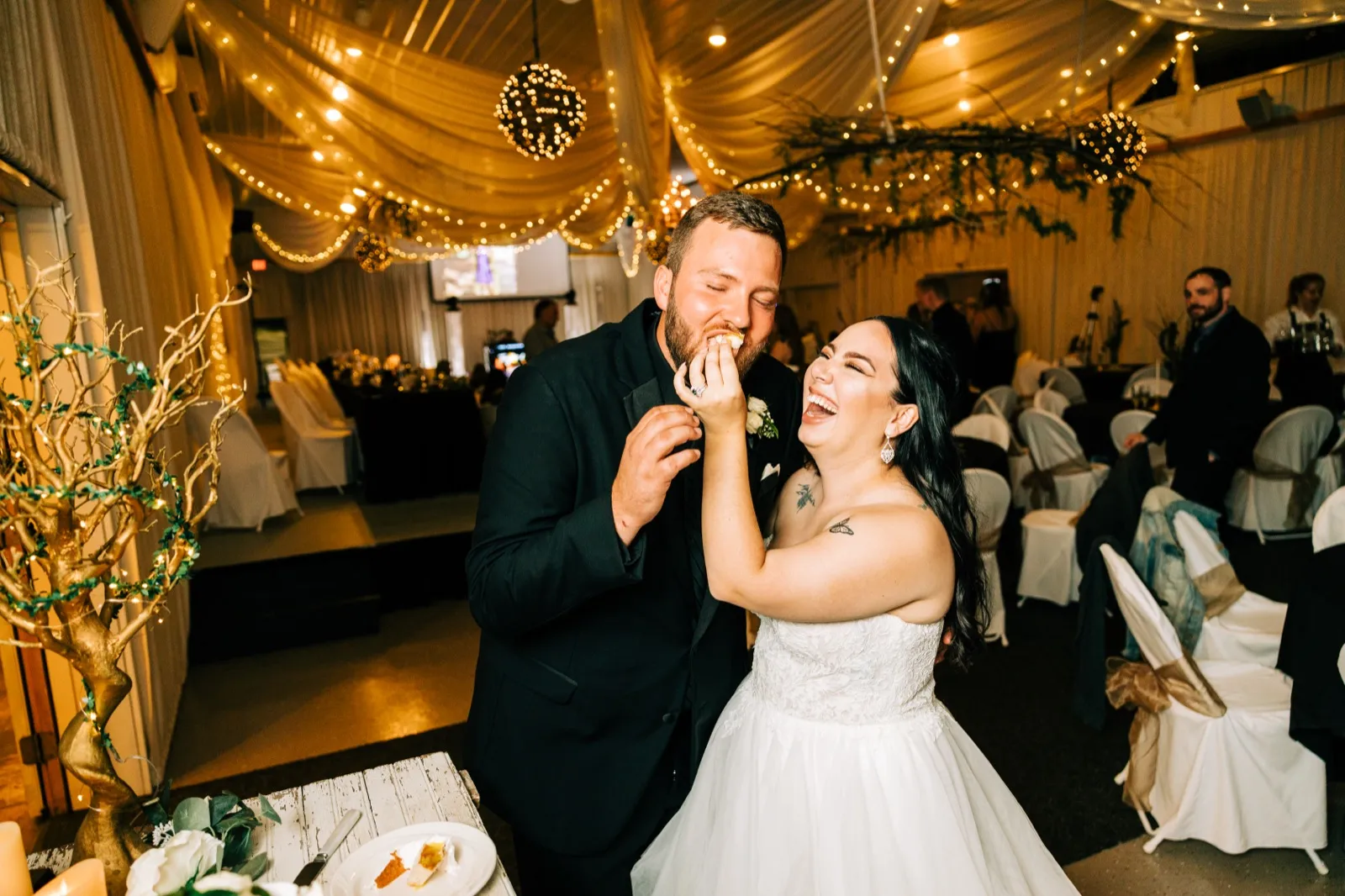 The bride laughs openly as she feeds the groom a bite of cake beneath draped fairy lights at Pine Peaks Event Center — Tim Larsen Photography, Brainerd Lakes MN