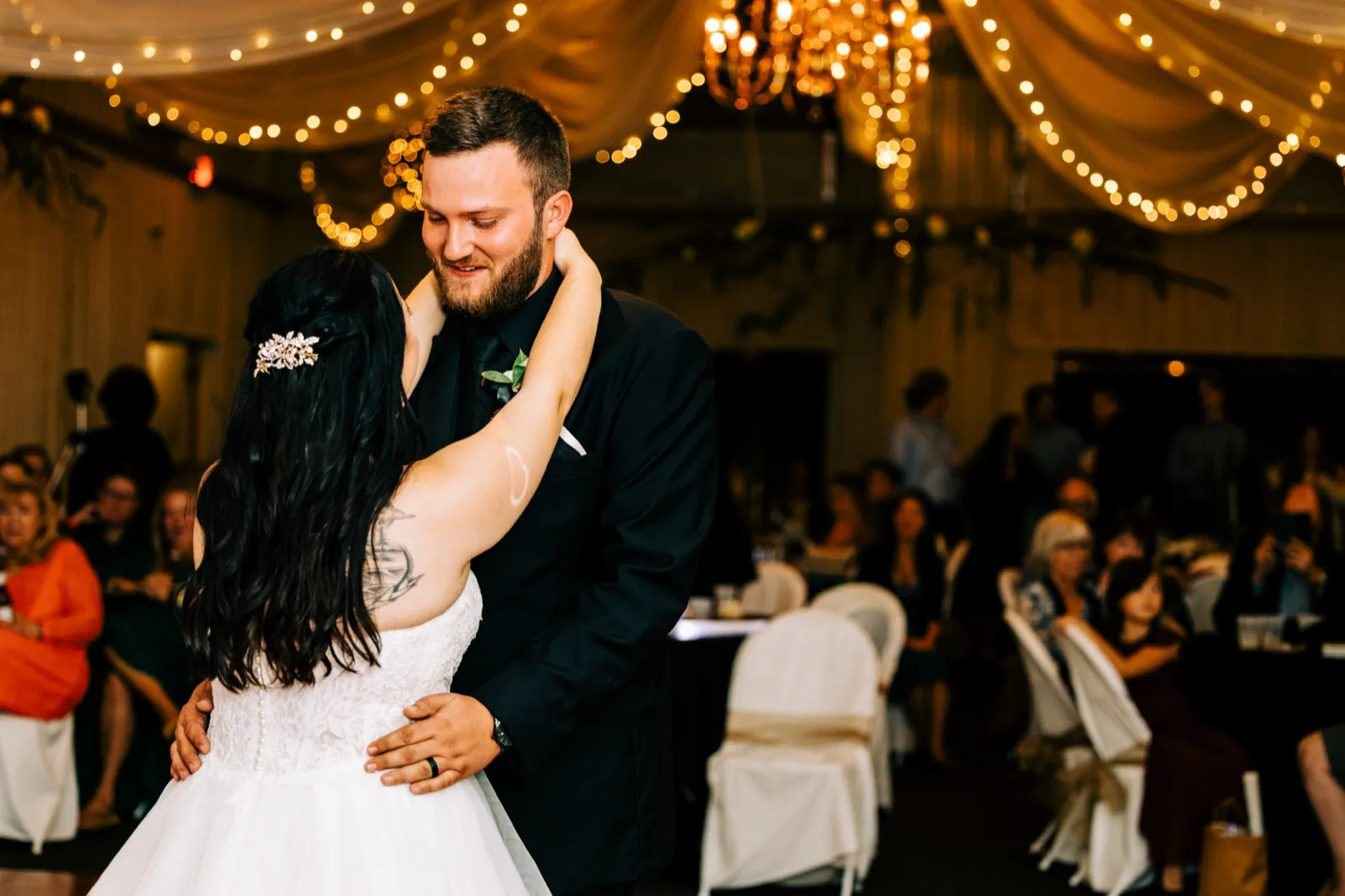 Charles smiles down at Taylor as they slow dance beneath draped fabric and chandelier lights at their reception, with seated guests watching in the background. — Tim Larsen Photography, Brainerd Lakes MN