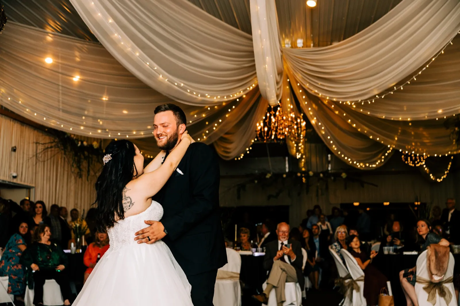 The bride and groom share their first dance beneath a ceiling of draped fabric and string lights at Pine Peaks Event Center, with seated guests watching from the edges of the room — Tim Larsen Photography, Brainerd Lakes MN