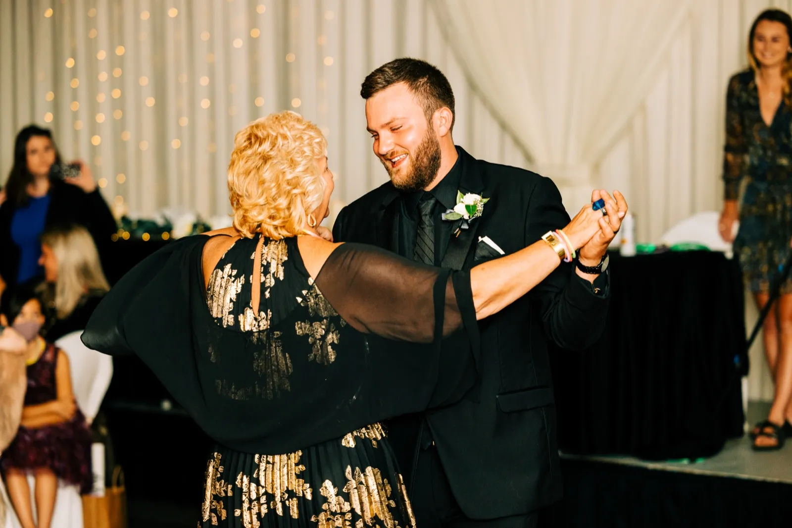 The groom smiles down at his mother as they dance together on a reception dance floor with a curtain of twinkle lights behind them — Tim Larsen Photography, Brainerd Lakes MN