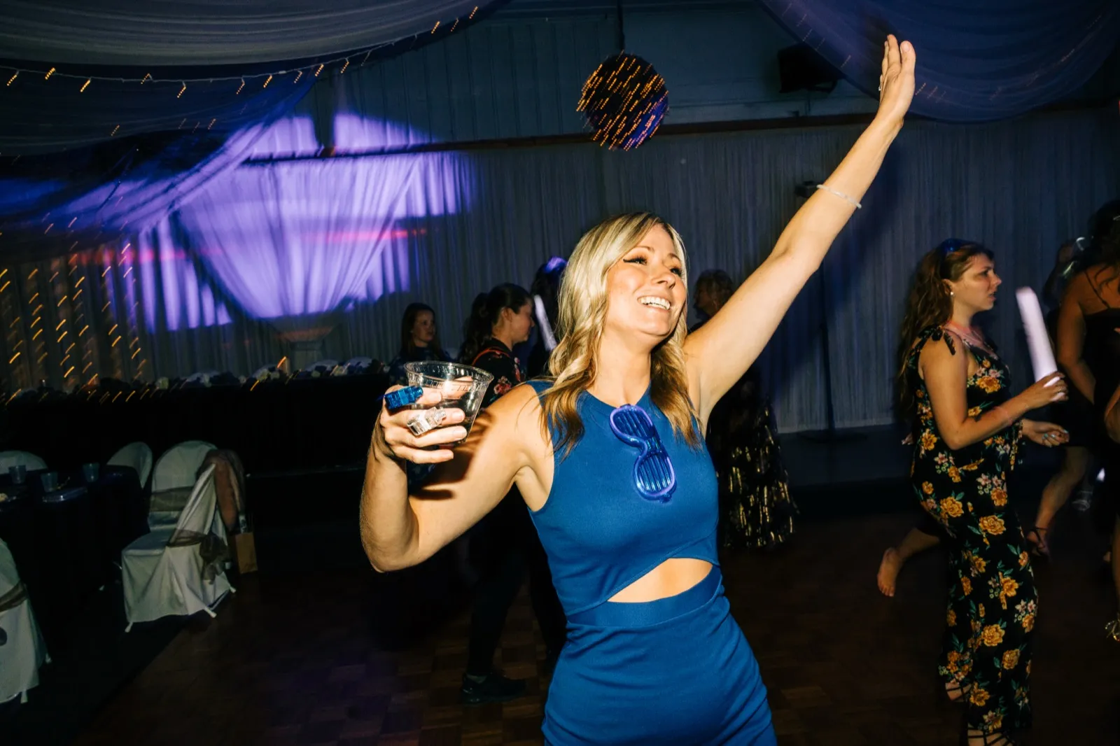 A blonde guest in a blue cutout dress laughs with her arm raised on the dance floor, holding a drink and wearing novelty shutter shades, with a disco ball and draped ceiling above. — Tim Larsen Photography, Brainerd Lakes MN
