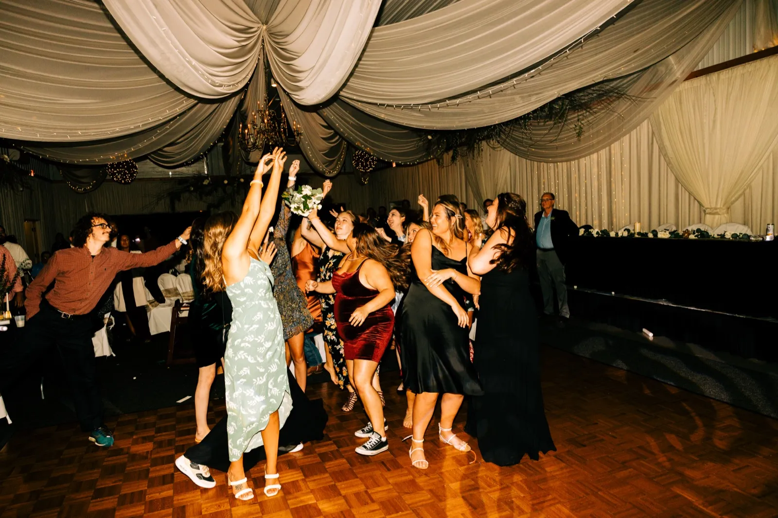Guests reach upward as Taylor's bouquet hangs midair beneath a draped ceiling and chandelier on the dance floor. — Tim Larsen Photography, Brainerd Lakes MN