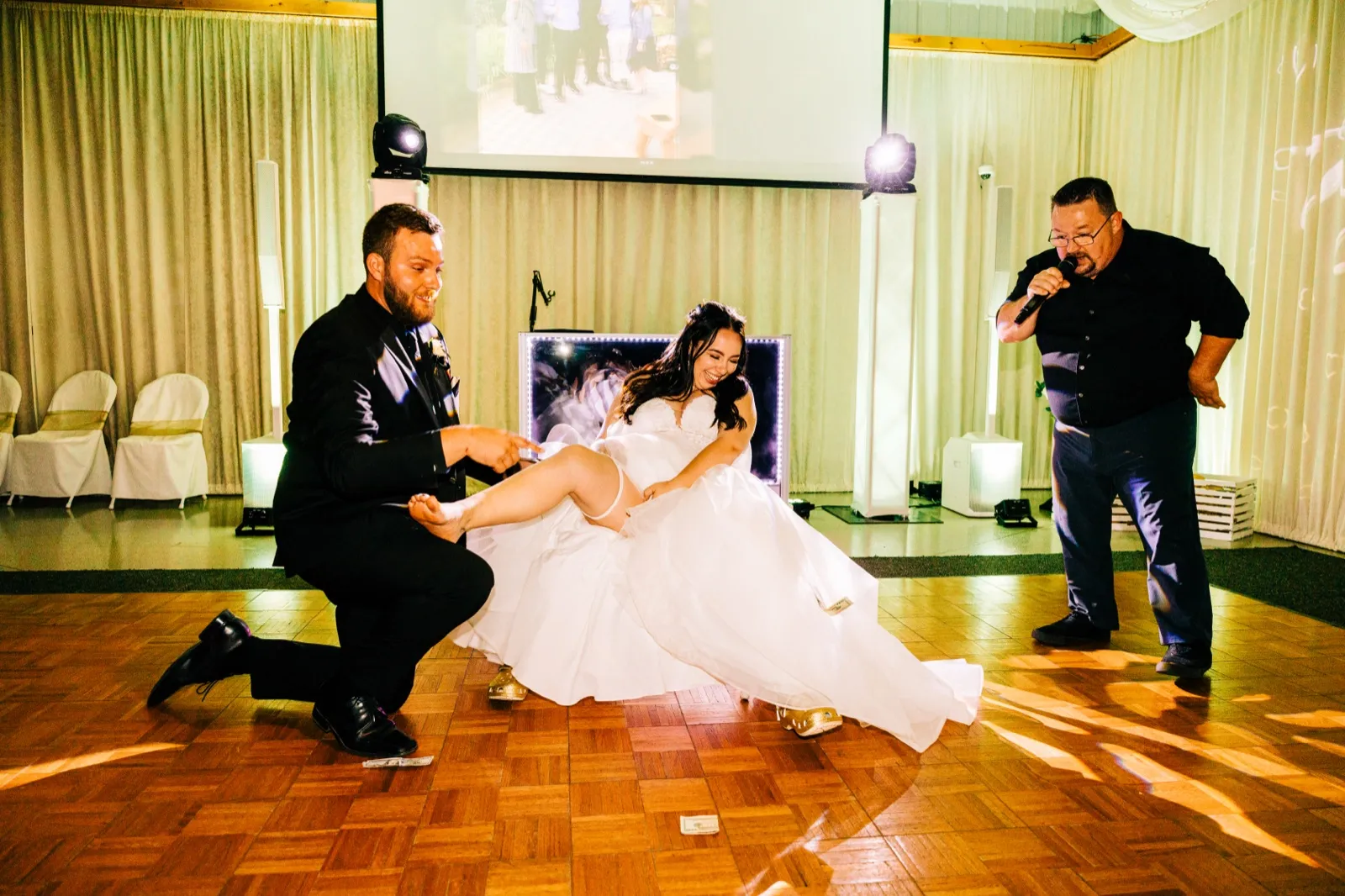 Charles kneels on the dance floor to remove Taylor's garter as she laughs and an emcee speaks into a microphone nearby. — Tim Larsen Photography, Brainerd Lakes MN