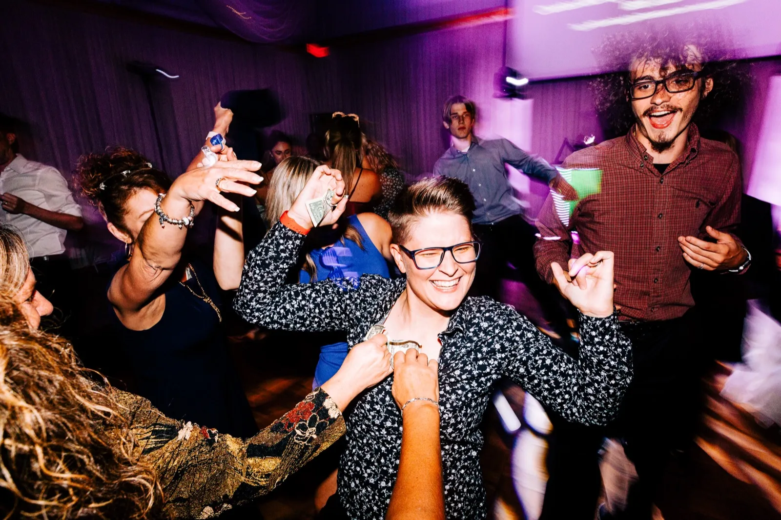 A grinning guest in glasses and a floral shirt dances at the center of a crowded dance floor lit purple, with dollar bills tucked into her collar and other guests dancing in motion-blurred energy around her. — Tim Larsen Photography, Brainerd Lakes MN