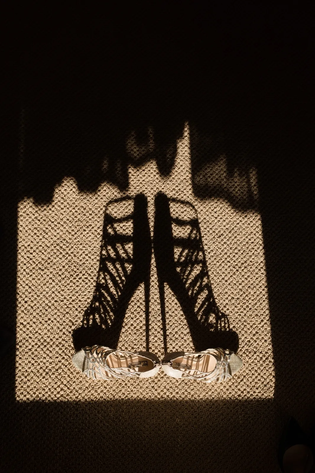 A pair of strappy silver bridal heels sits in a beam of window light on textured carpet, casting elongated shadows. — Tim Larsen Photography, Brainerd Lakes MN