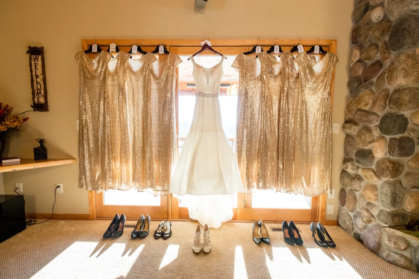 The bride's white wedding dress hangs in a sunlit window flanked by six gold sequined bridesmaid dresses, with the bridal party's shoes lined up on the carpet below. — Tim Larsen Photography, Brainerd Lakes MN