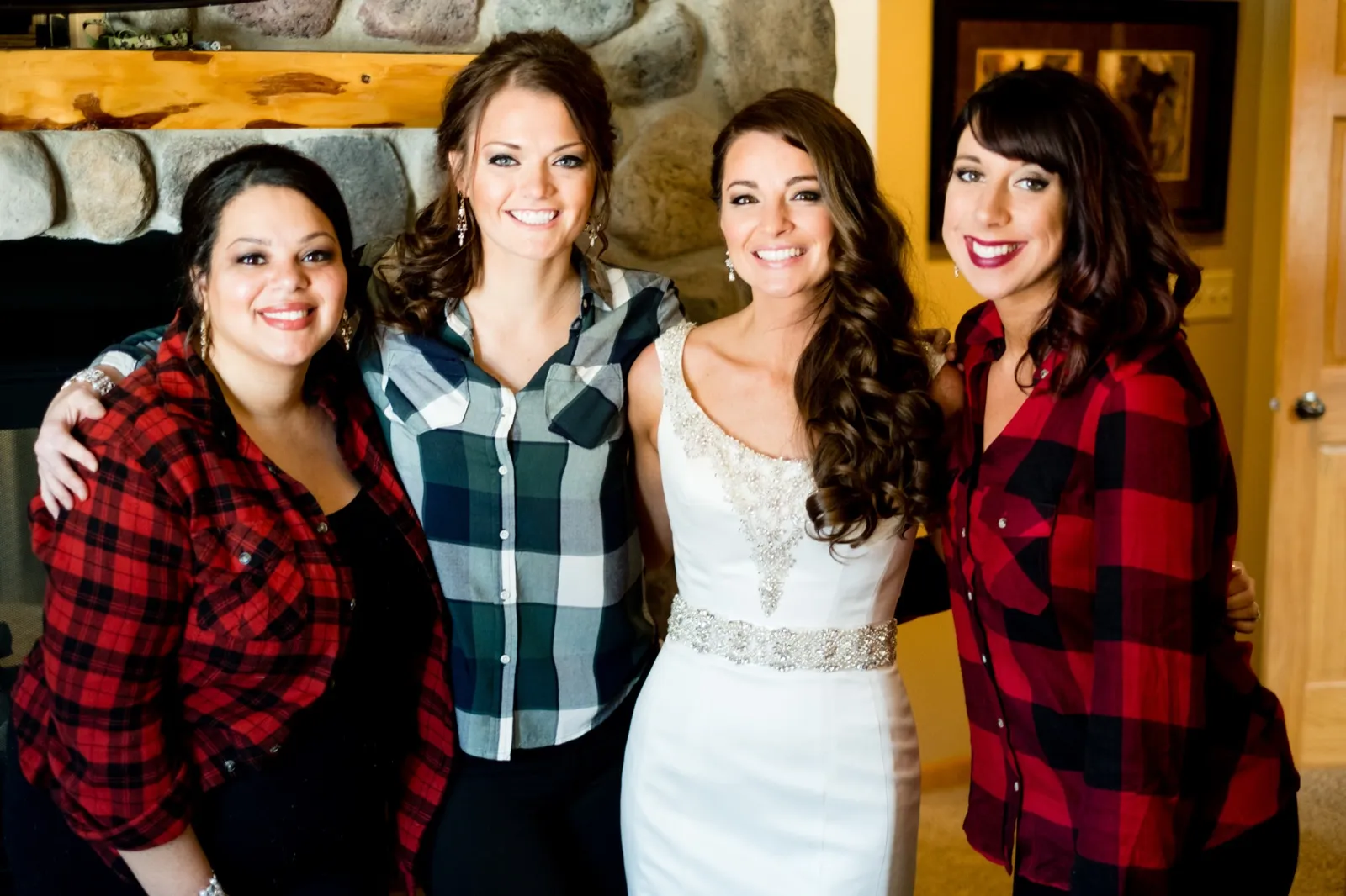 The bride in a beaded white gown stands smiling with three friends in flannel shirts in front of a stone fireplace. — Tim Larsen Photography, Brainerd Lakes MN
