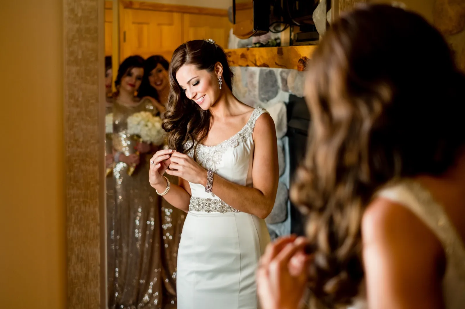 The bride smiles down at her ring while standing in a doorway, with two bridesmaids in gold dresses visible in a mirror reflection behind her. — Tim Larsen Photography, Brainerd Lakes MN