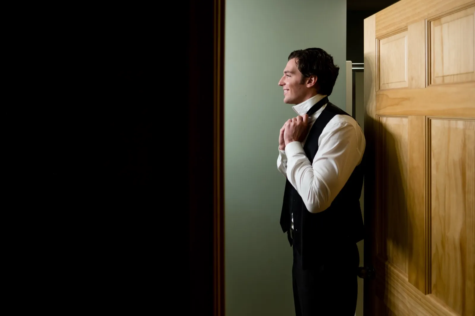 The groom stands in a doorway adjusting his bow tie, lit by soft window light, with a large area of dark wall to his left. — Tim Larsen Photography, Brainerd Lakes MN