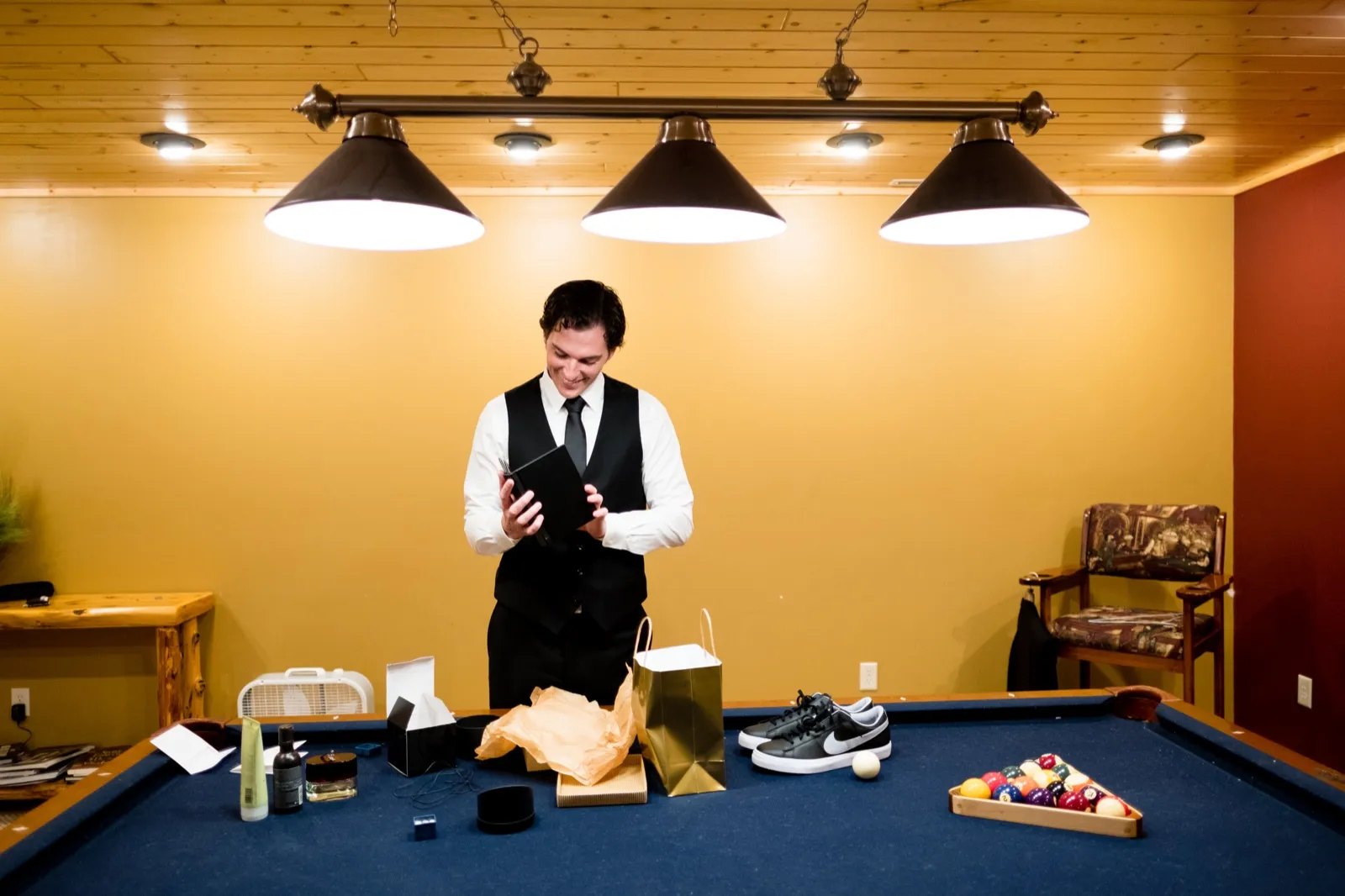 The groom in a black vest and tie smiles as he reads a card while standing behind a pool table covered with opened gifts beneath three pendant lights. — Tim Larsen Photography, Brainerd Lakes MN