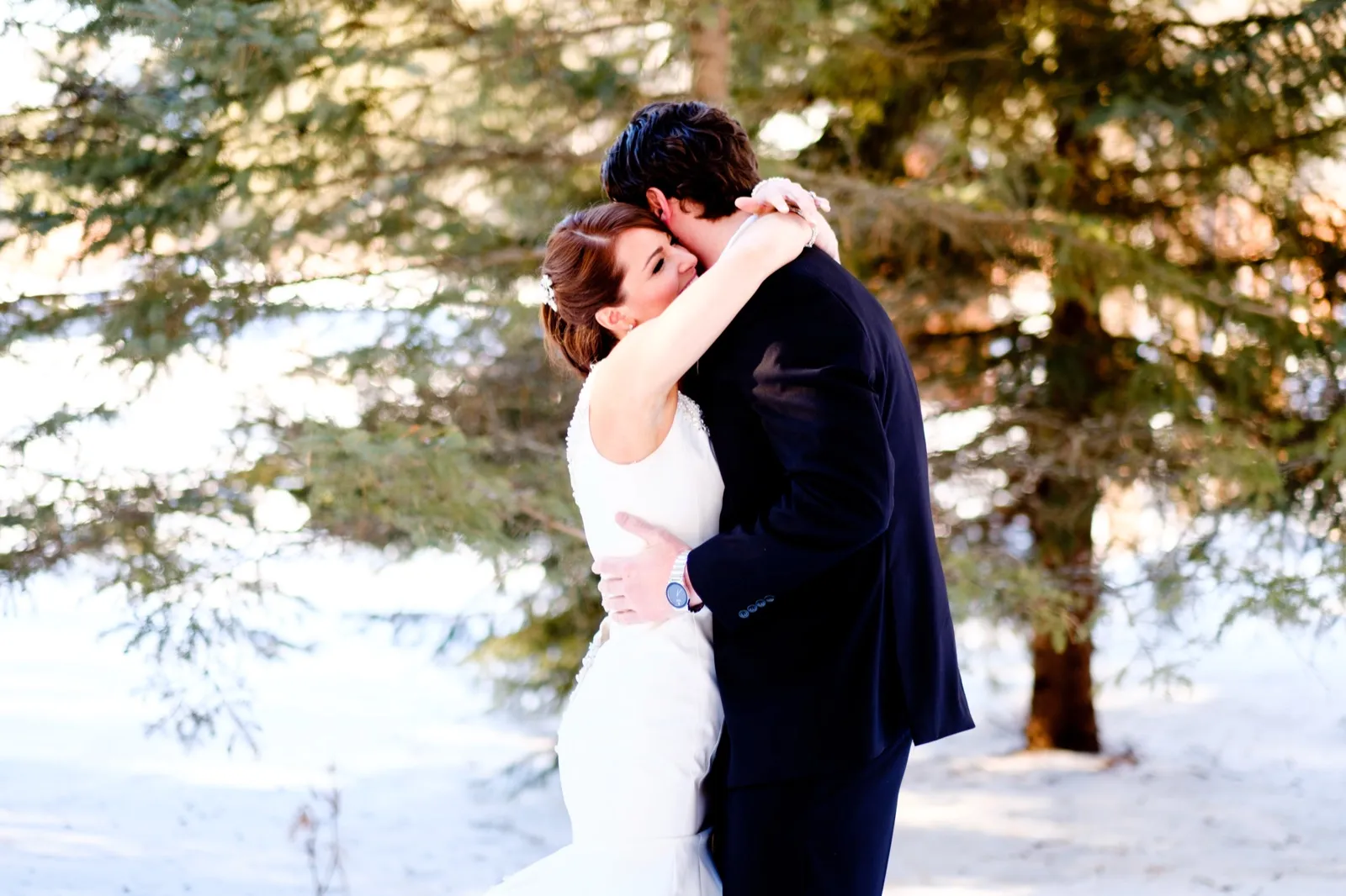 The bride in a sleeveless white gown presses her face into the groom's neck as they embrace among sunlit evergreens in the snow. — Tim Larsen Photography, Brainerd Lakes MN