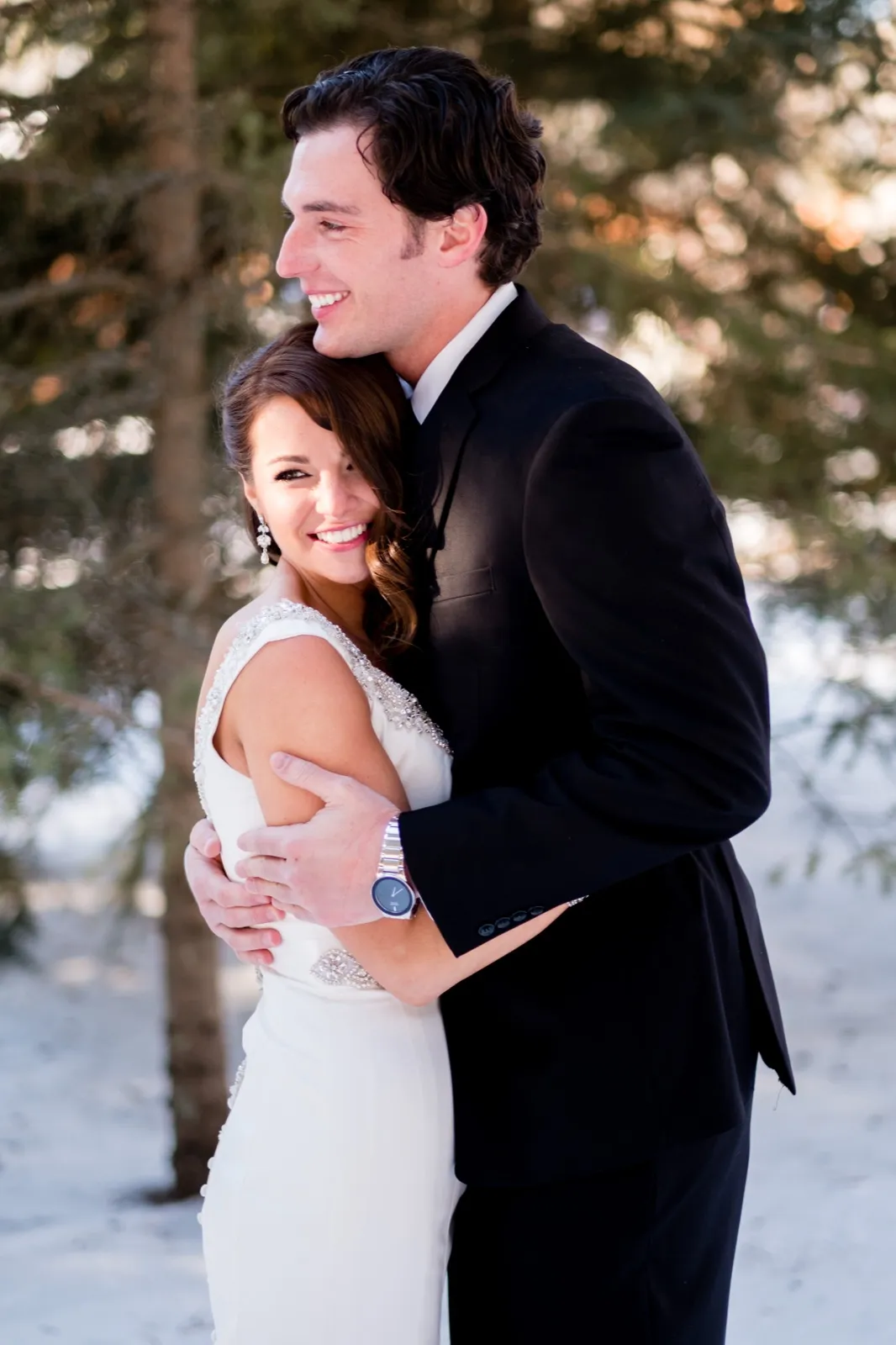 The bride smiles into the camera while the groom holds her close in a snowy evergreen grove during a winter portrait session. — Tim Larsen Photography, Brainerd Lakes MN