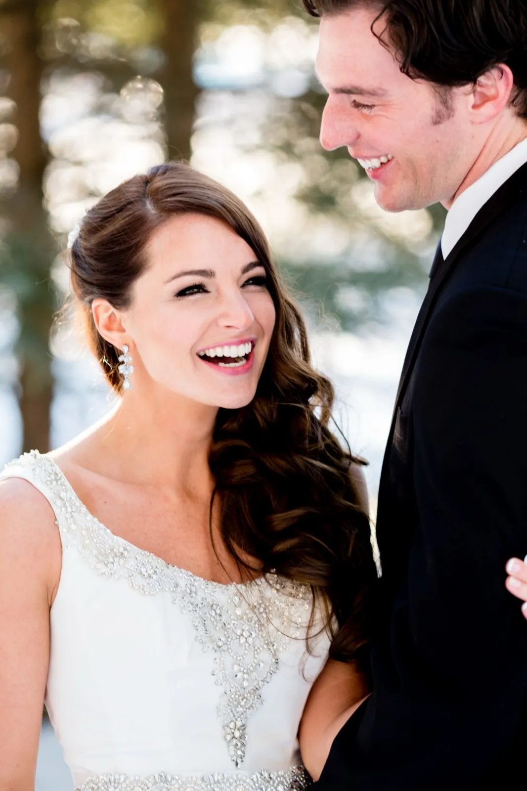 The bride laughs openly while the groom gazes at her with a smile, both backlit by winter sun in a snowy pine forest. — Tim Larsen Photography, Brainerd Lakes MN