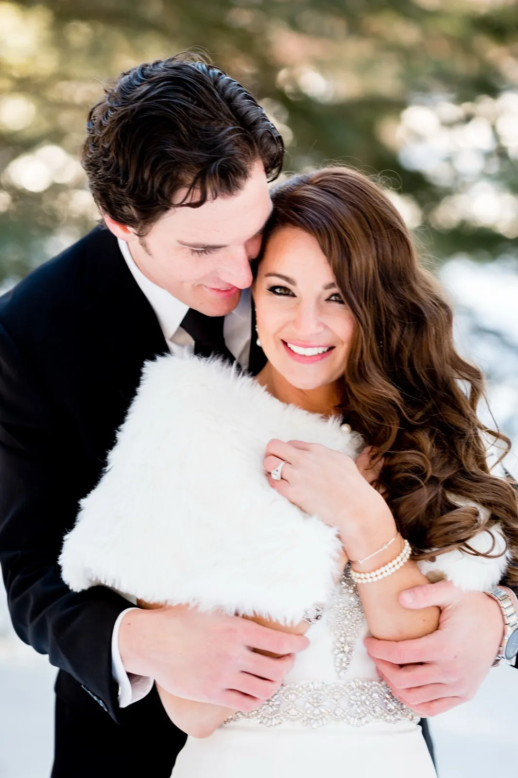 The groom nuzzles into the bride's hair as she smiles at the camera, wrapped in a white fur stole and beaded gown in a snowy pine grove. — Tim Larsen Photography, Brainerd Lakes MN