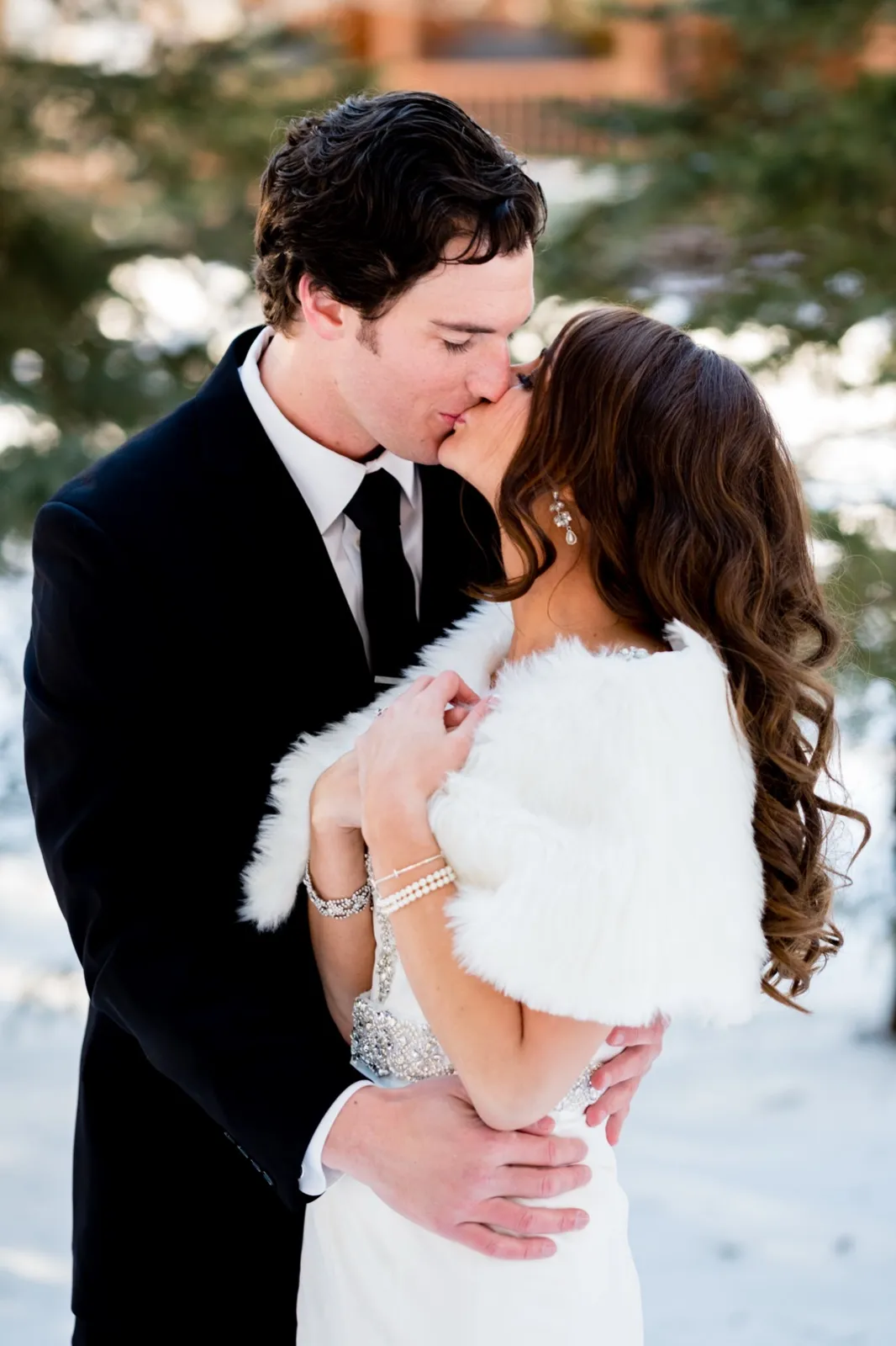 The bride in a white fur stole and beaded gown kisses the groom in a dark suit as they stand together in the snow. — Tim Larsen Photography, Brainerd Lakes MN