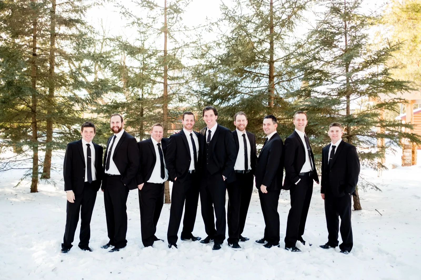 The groom and his groomsmen in black tuxedos stand in a diagonal line on snow-covered ground surrounded by tall pine trees. — Tim Larsen Photography, Brainerd Lakes MN