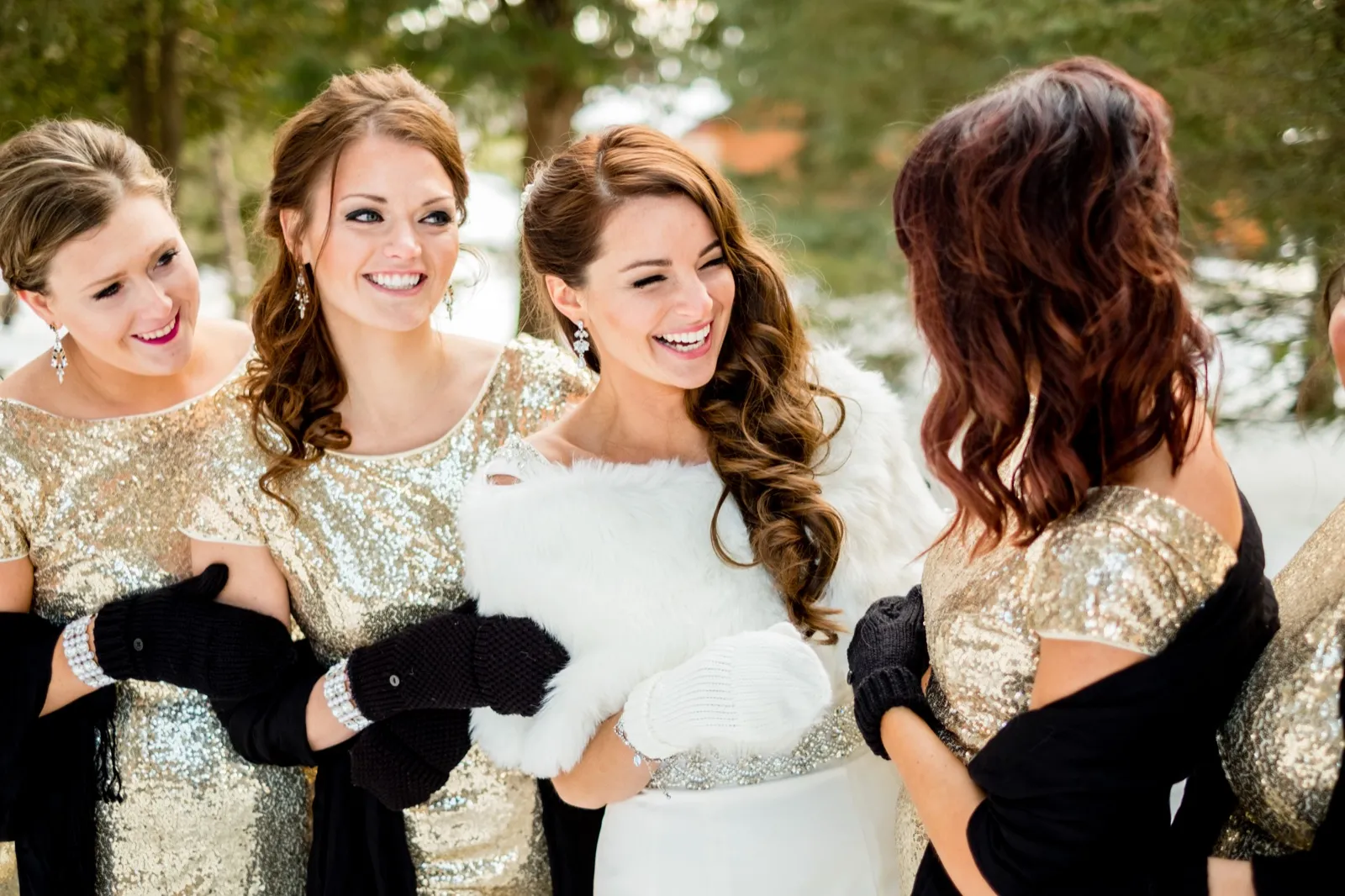 The bride laughs in a white fur wrap surrounded by her bridesmaids in gold sequin dresses and black mittens in a snowy evergreen grove. — Tim Larsen Photography, Brainerd Lakes MN