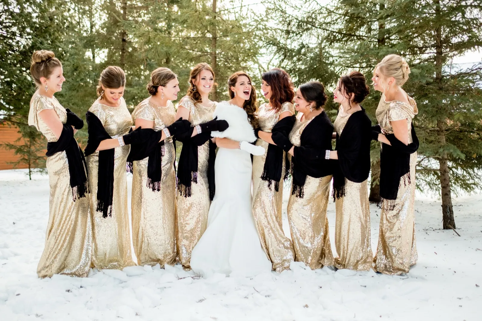 The bride laughs open-mouthed at the center of her bridal party, all wearing gold sequin gowns and black wraps, standing in a snowy clearing surrounded by pine trees. — Tim Larsen Photography, Brainerd Lakes MN