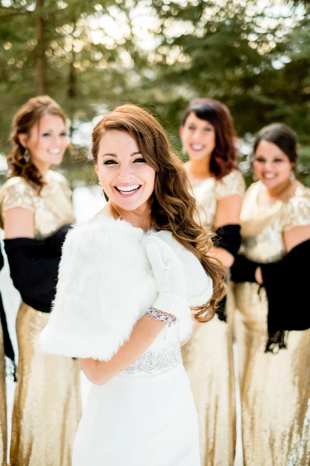 The bride smiles brightly while holding a white fur wrap, with three bridesmaids in gold dresses softly out of focus behind her in a snowy forest. — Tim Larsen Photography, Brainerd Lakes MN