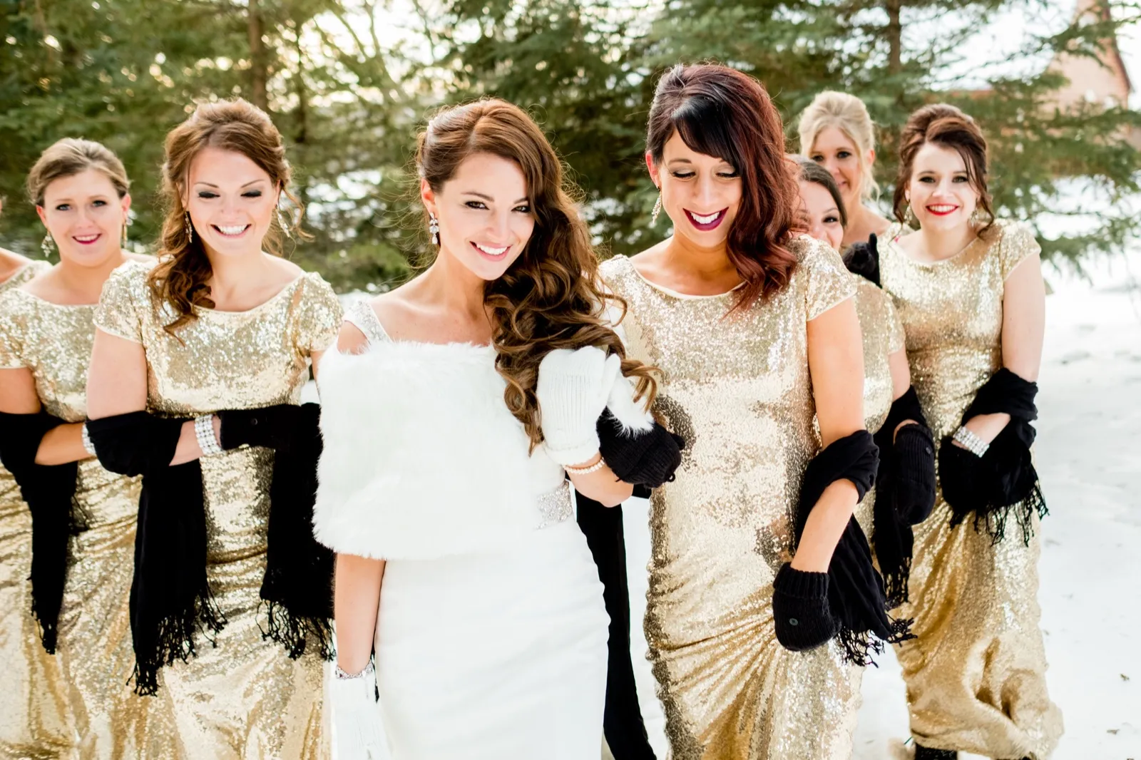 The bride in a white fur stole walks through snow with six bridesmaids in gold sequin gowns and black shawls, all smiling and looking down. — Tim Larsen Photography, Brainerd Lakes MN