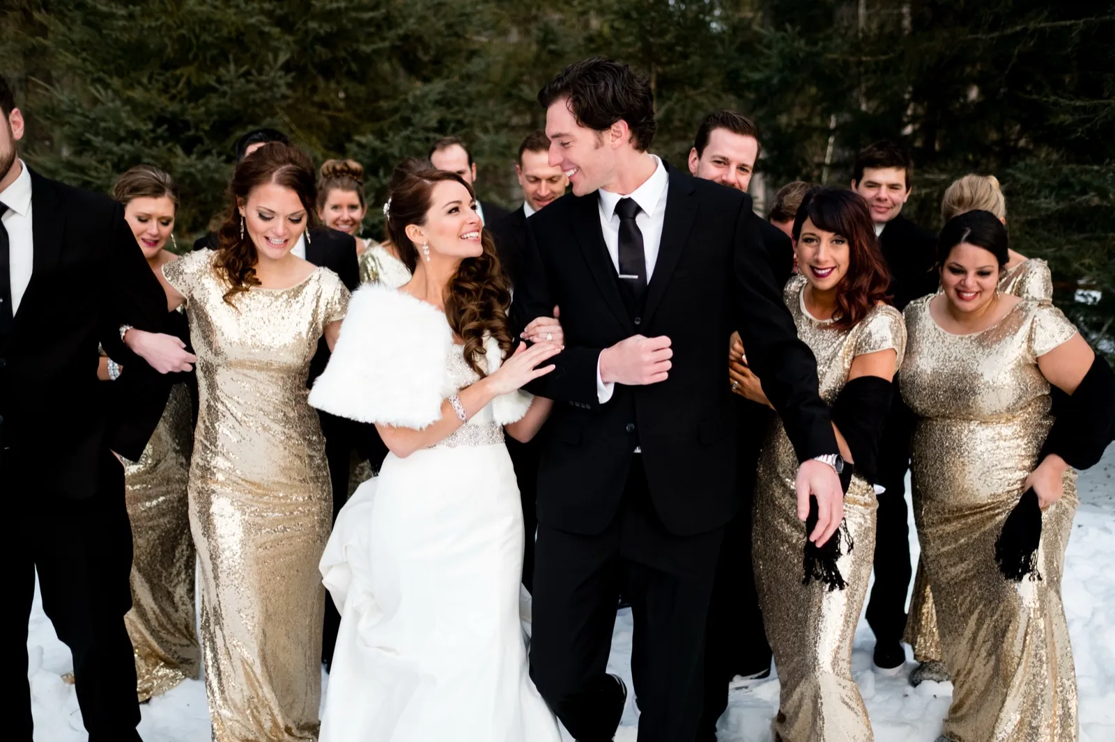 The bride and groom laugh together at the center of their wedding party, with bridesmaids in gold sequined gowns and groomsmen in black suits standing in fresh snow before a wall of evergreens. — Tim Larsen Photography, Brainerd Lakes MN